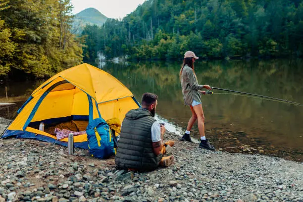 Angler casting a fishing rod into a serene lake surrounded by lush greenery, equipped with essential fishing gear.